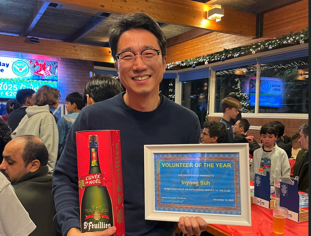 A smiling person holding a gift box of beer and an award certificate labeled 'Volunteer of the Year' in a festive gathering with blurred attendees in the background.